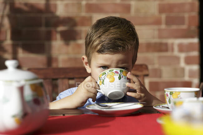 Niño con el pelo castaño sentado al aire libre en una mesa, bebiendo de la taza. - foto de stock
