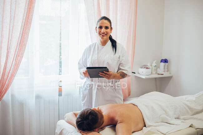 Estetista sorridente in piedi accanto alla donna sdraiata sul letto di trattamento nel salone di bellezza. — Foto stock