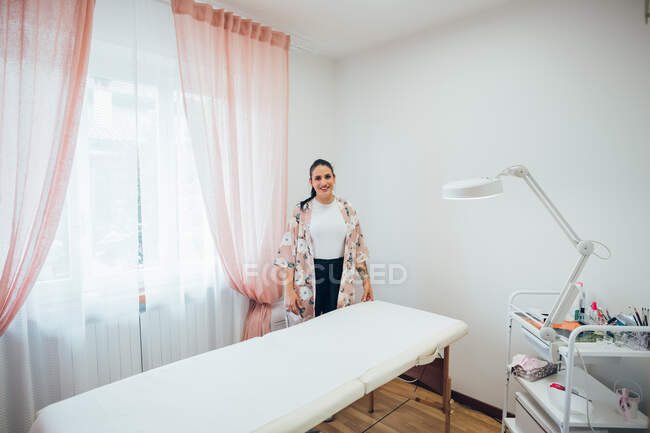 Косметолог стоїть поруч з ліжком для лікування в салоні краси, посміхаючись на камеру . — стокове фото