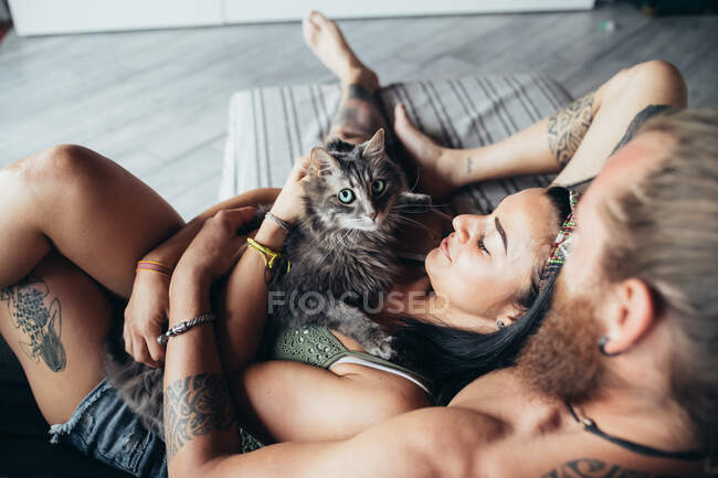 Bearded tattooed man with long brunette hair and woman with long brown hair cuddling with fluffy grey cat on a sofa. — Stock Photo