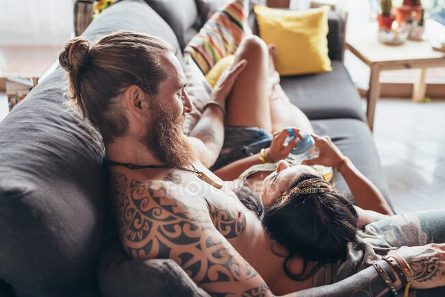 Bärtiger tätowierter Mann mit langen brünetten Haaren und Frau mit langen braunen Haaren kuschelt auf einem Sofa. — Stockfoto