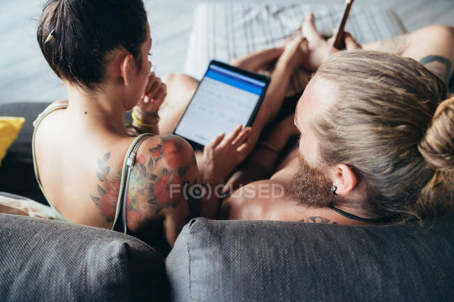 Bearded tattooed man with long brunette hair and woman with long brown hair sitting on a sofa, looking at digital tablet. — Stock Photo