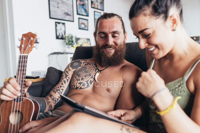 Бородатий татуйований чоловік з довгим брюнетка волоссям і жінка з довгим коричневим волоссям сидить на дивані, дивлячись на цифровий планшет . — стокове фото