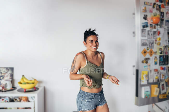 Smiling woman with long brown hair wearing vest top and hot pants standing in a kitchen. — Stock Photo