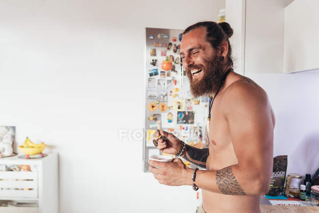 Bearded tattooed shirtless man with long brunette hair standing in a kitchen, laughing. — Stock Photo