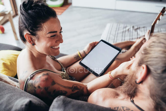 Hombre tatuado barbudo con el pelo largo morena y mujer con el pelo largo marrón sentado en un sofá, mirando la tableta digital. - foto de stock