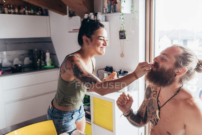 Bearded tattooed man with long brunette hair and woman with long brown hair in a kitchen, feeding each other. — Stock Photo