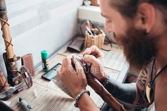 Bearded tattooed man with long brunette hair sitting at table, holding axe with wood and leather handle. — Stock Photo