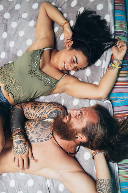 Bärtiger tätowierter Mann mit langen brünetten Haaren und Frau mit langen braunen Haaren liegen auf einem Bett und lächeln einander an. — Stockfoto