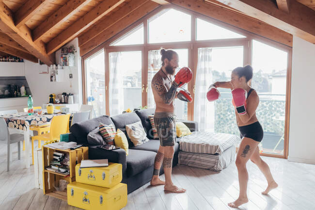 Homme tatoué barbu aux longs cheveux bruns et femme aux longs cheveux bruns debout à l'intérieur, pratiquant le kickboxing. — Photo de stock