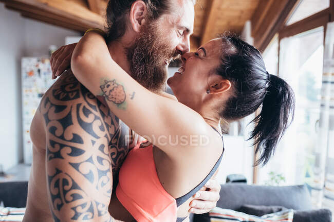 Barbudo hombre tatuado con el pelo largo morena y mujer con el pelo largo marrón de pie en el interior, abrazos y besos. - foto de stock