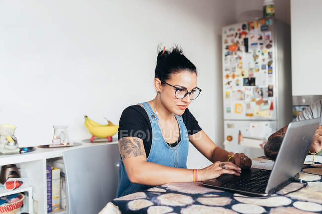 Жінка з довгим коричневим волоссям в окулярах сидить за кухонним столом, використовуючи ноутбук . — стокове фото