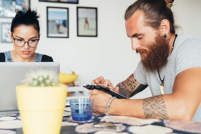 Bearded tattooed man with long brunette hair and woman with long brown hair sitting at a kitchen table, using laptop and mobile phone. — Stock Photo