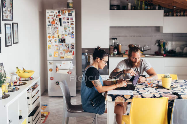 Homem tatuado barbudo com cabelos longos morena e mulher com cabelos longos marrom sentado em uma mesa de cozinha, usando laptop e telefone celular. — Fotografia de Stock
