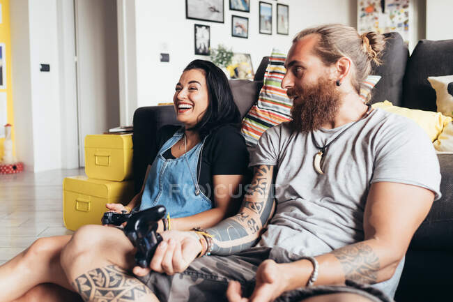 Barbudo hombre tatuado con el pelo largo morena y la mujer con el pelo largo marrón sentado en un sofá, sonriendo mientras juega juego de consola. - foto de stock