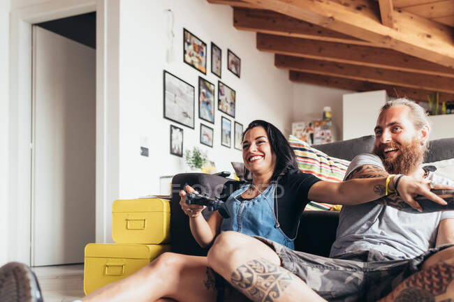 Barbudo hombre tatuado con el pelo largo morena y la mujer con el pelo largo marrón sentado en un sofá, sonriendo mientras juega juego de consola. - foto de stock