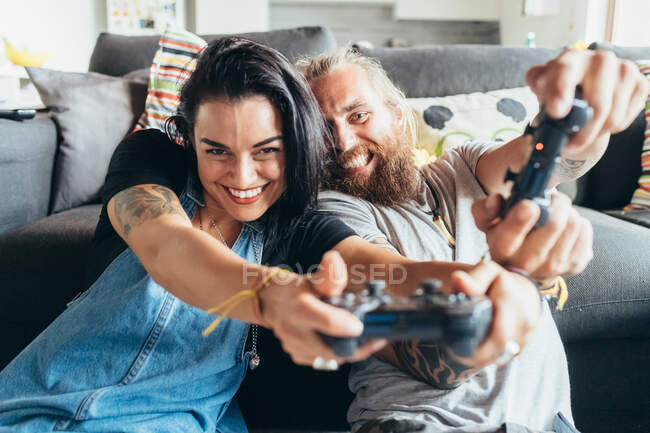 Бородатий татуйований чоловік з довгим волоссям брюнетки і жінка з довгим коричневим волоссям сидить на дивані, посміхаючись, граючи в консольну гру . — стокове фото
