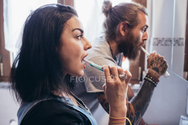 Мужчина с татуировкой на бородке с длинными брюнетками и женщина с длинными каштановыми волосами, стоящими перед зеркалом, чистящие зубы. — стоковое фото