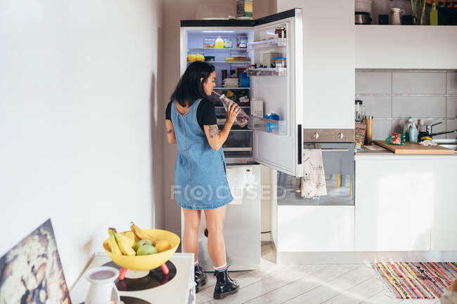 Вид спереду татуйованої жінки з довгим коричневим волоссям у джинсовій сукні, що стоїть перед відкритим холодильником, п'є з пляшки . — стокове фото