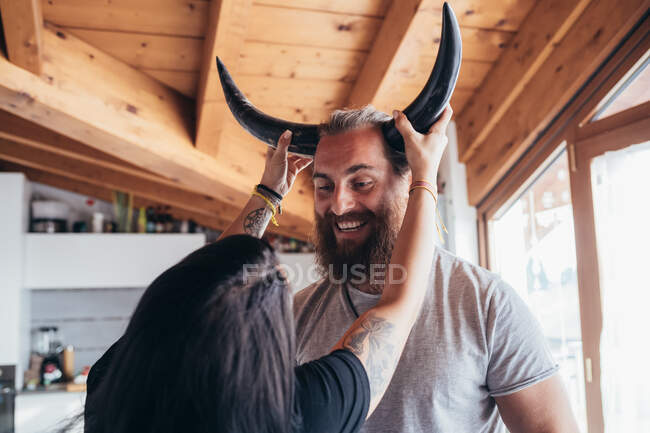 Mulher segurando chifres de vaca na cabeça de homem barbudo sorridente com cabelo morena. — Fotografia de Stock