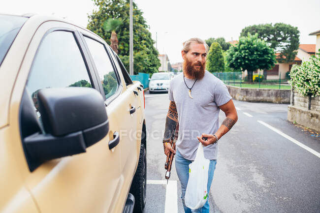 Мужчина с татуировкой на бородке с длинными волосами брюнетки с пластиковыми пакетами для покупок, идущий к пикапу. — стоковое фото