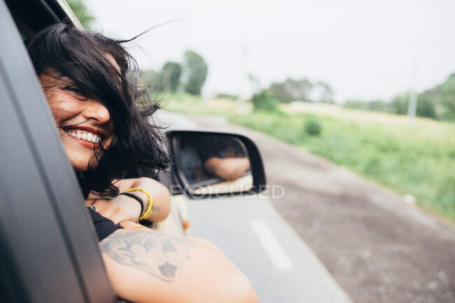 Smiling woman with long brown hair and tattoos looking out of car window. — Stock Photo