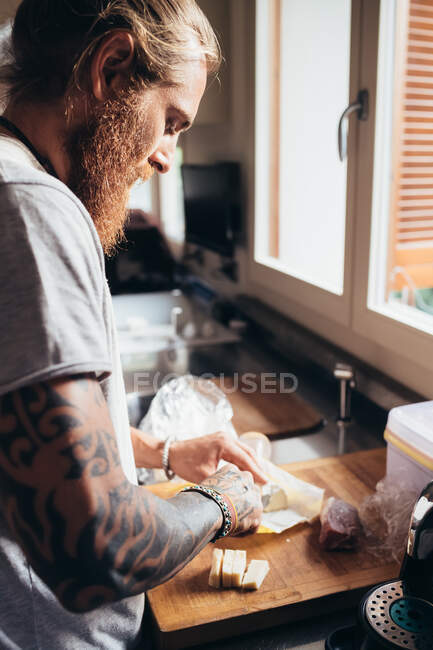 Bärtiger tätowierter Mann mit langen brünetten Haaren steht in einer Küche und bereitet Essen zu. — Stockfoto