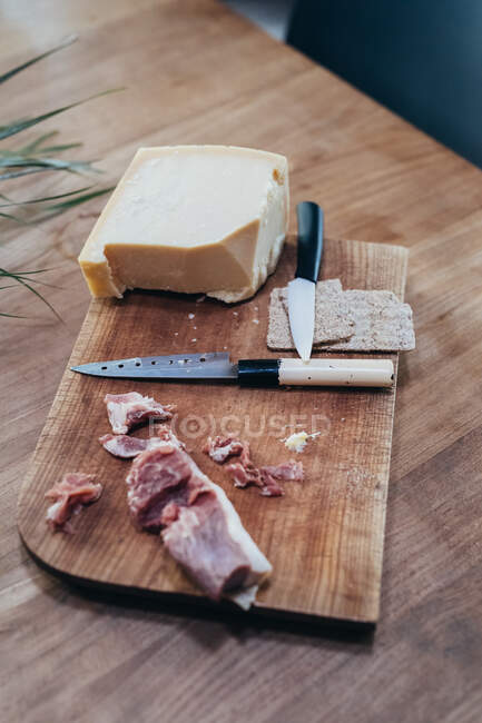 Alto angolo primo piano di coltelli, prosciutto affumicato e formaggio duro sul tagliere di legno. — Foto stock