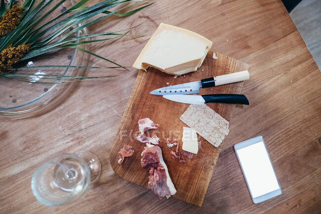 High angle close up of knives, smoked ham and hard cheese on wooden cutting board. — Stock Photo