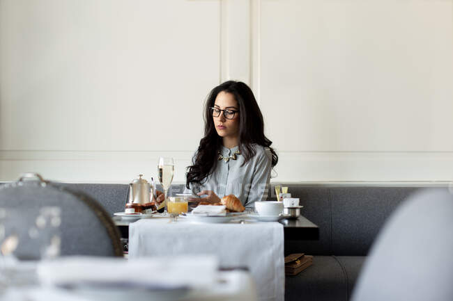 Donna con lunghi capelli neri che indossa occhiali seduti al tavolo di un ristorante, utilizzando il telefono cellulare. — Foto stock