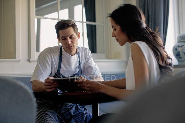 Chef vistiendo delantal azul y mujer sentada en una mesa, mirando la tableta digital. - foto de stock