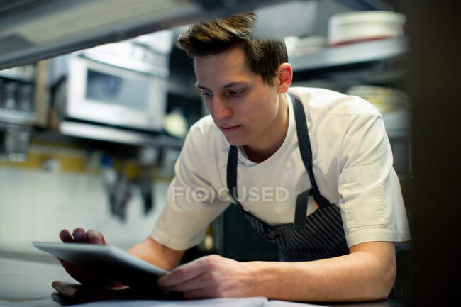 Chef wearing blue apron standing in kitchen, using digital tablet. — Stock Photo