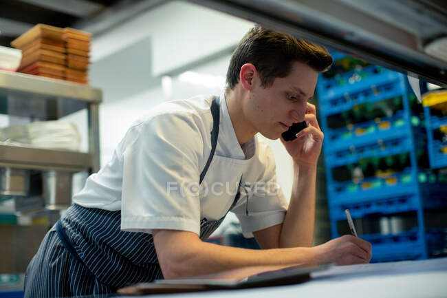 Chef vistiendo delantal azul de pie en la cocina, usando el teléfono móvil. - foto de stock