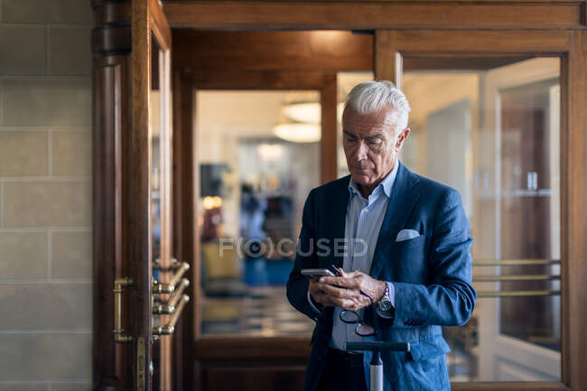 Старший бізнесмен стоїть в готельному лобі, дивлячись на мобільний телефон . — стокове фото