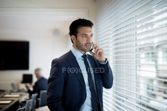 Homem de negócios barbudo vestindo terno e gravata em pé por uma janela, usando telefone celular. — Fotografia de Stock