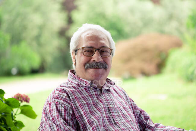 Porträt eines älteren Mannes mit Schnurrbart und Brille, der im Garten sitzt und in die Kamera lächelt. — Stockfoto