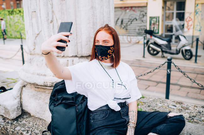 Молода жінка носить маску для обличчя під час вірусу Корони, сидячи на відкритому повітрі, приймаючи селфі . — стокове фото