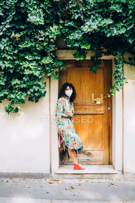 Junge Frau mit Gesichtsmaske während Coronavirus, steht vor Haustür und blickt in Kamera. — Stockfoto