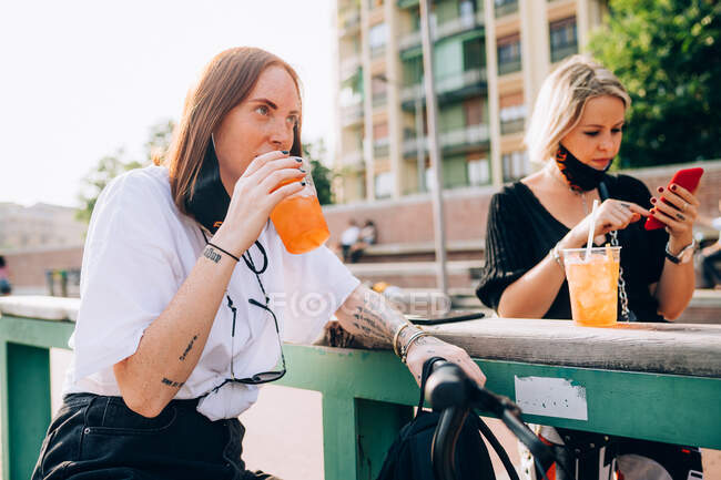 Due giovani donne sedute all'aperto su una panchina, che bevono e usano il cellulare. — Foto stock