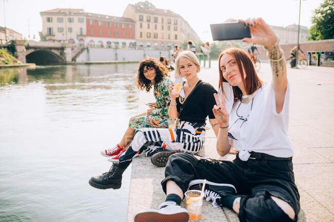 Três jovens mulheres sentadas em uma margem do rio, tirando selfie. — Fotografia de Stock