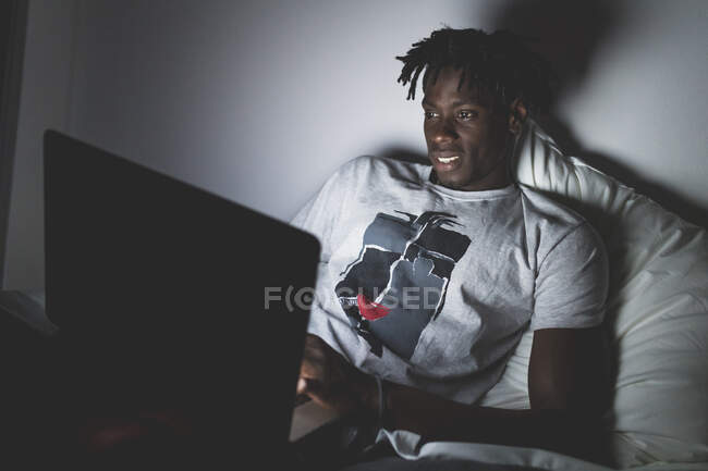 Junger Mann mit kurzen Dreadlocks liegt nachts im Bett und schaut auf Laptop. — Stockfoto