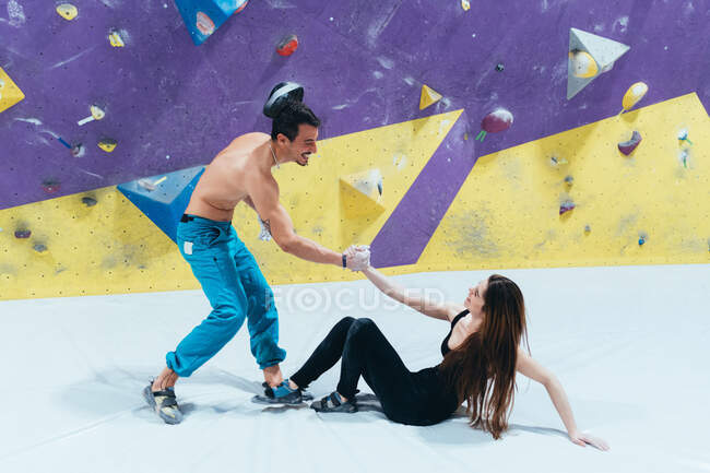 Man helping up woman sitting on floor in front of indoor rock climbing wall. — Stock Photo