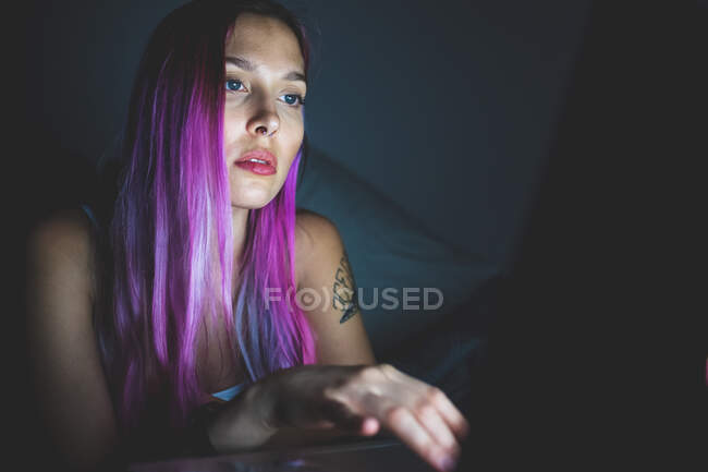 Mujer joven con el pelo largo y rosa mirando a un ordenador portátil, la cara iluminada por el resplandor de la pantalla. - foto de stock