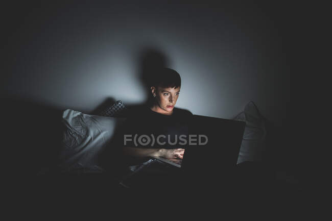 Jovem com cabelo curto deitado na cama à noite, olhando para laptop. — Fotografia de Stock