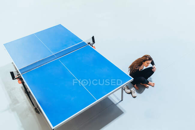 Vista de ángulo alto de la mujer sentada en el suelo descalza junto a la mesa de ping pong azul - foto de stock