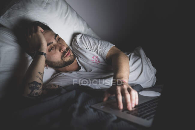 Jovem barbudo deitado na cama à noite, olhando para monitor de tela do laptop — Fotografia de Stock