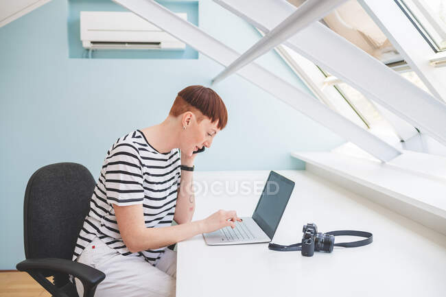 Молода жінка з коротким волоссям сидить за столом, дивлячись на ноутбук — стокове фото