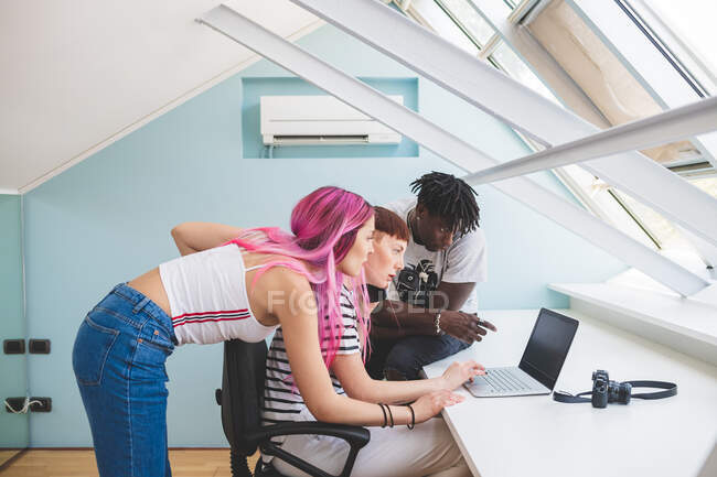 Dos mujeres jóvenes y un hombre joven sentado en el escritorio, mirando la pantalla del ordenador portátil - foto de stock