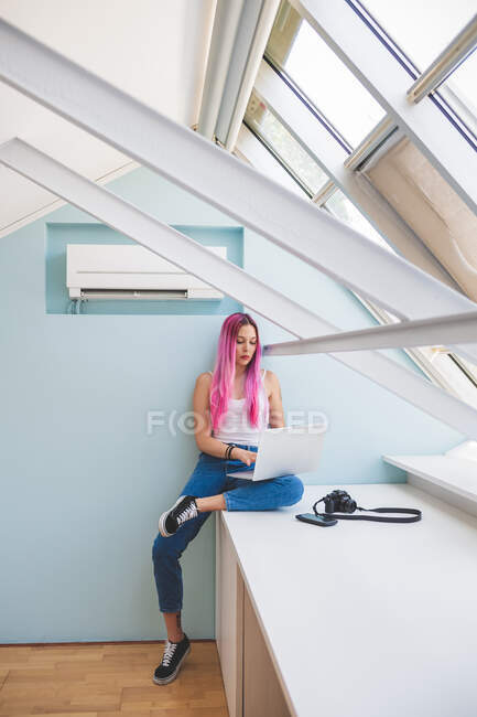 Молода жінка з довгим рожевим волоссям сидить на столі і друкує на ноутбуку. — стокове фото