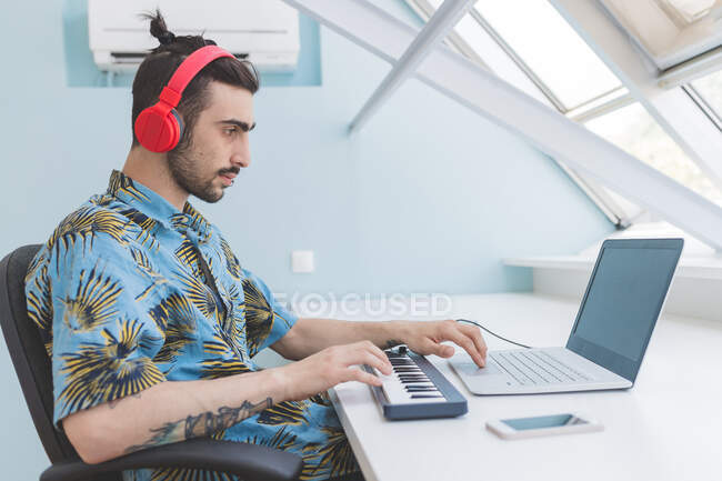 Молодой бородатый мужчина с пирсингом сидит за столом, в красных наушниках, печатает на ноутбуке и клавиатуре — стоковое фото